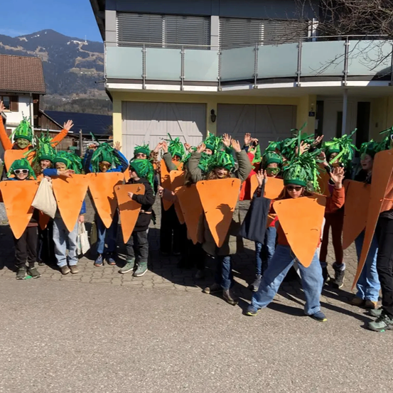 Eine Gruppe von Menschen in Karottenkostümen posiert für ein Foto vor einem Gebäude mit Bergen im Hintergrund.
