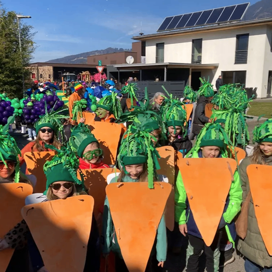 Eine Gruppe von Kindern in Karottenkostümen steht in einer Reihe im Freien. Sie lächeln und schauen in die Kamera. Im Hintergrund sind Gebäude und Berge zu sehen.