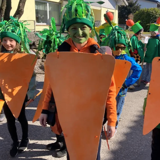 Eine Gruppe von Kindern in Möhrenkostümen geht die Straße entlang. Jedes Kind hält eine große orange Möhre in der Hand.