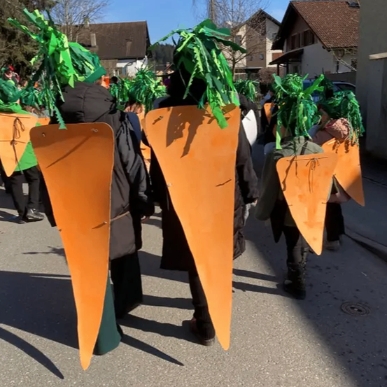 Eine Gruppe von Kindern in Karottenkostümen, die orangefarbene karottenförmige Requisiten halten, laufen bei einem Umzug auf einer Straße mit Häusern im Hintergrund.