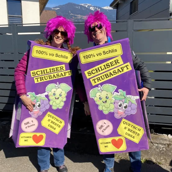 Zwei Personen in Kostümen, die in Form von lila Boxen von Schliser Trubasaft gekleidet sind, stehen vor einem Haus mit Bergen im Hintergrund.