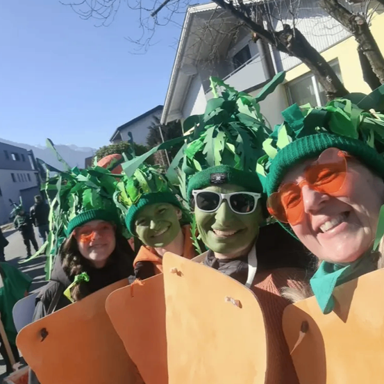 Vier Menschen, die als Karotten verkleidet sind, mit grünen Blättern auf den Köpfen und orangefarbenen Gläsern lächeln in die Kamera. Sie stehen vor Gebäuden und Bäumen an einem sonnigen Tag.