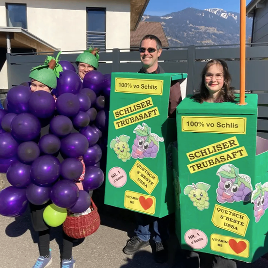 Vier Personen in Traubenkostümen und zwei grünen Schachteln mit Etiketten, eine davon mit der Aufschrift 'SCHLISER TRUBASAF 100% vo Schlis', stehen draußen. Dahinter befindet sich ein Gebäude mit einem Zaun, Bergen in der Ferne und einem klaren blauen Himmel.