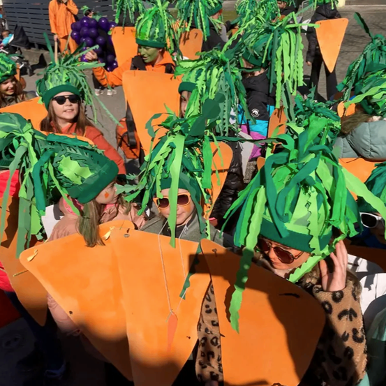 Eine Gruppe von Menschen in Karottenkostümen nimmt an einem Umzug teil, mit grünen Blättern auf dem Kopf und orangefarbenen Kostümen.