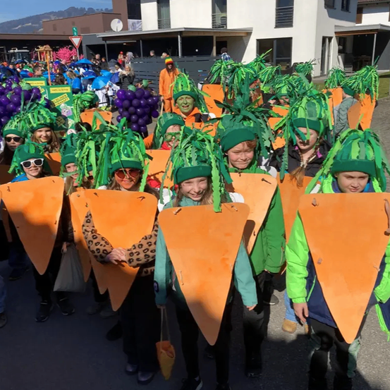 Eine Gruppe von Menschen, die als Karotten verkleidet sind, nehmen an einem Umzug teil. Sie tragen grüne Hüte und orange Kostüme. Einige halten Taschen. Gebäude und Menschen sind im Hintergrund.