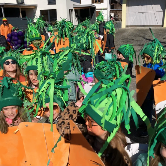 Eine Gruppe von Kindern in grünen und orangefarbenen Kostümen und Mützen versammelt sich im Freien. Einige halten orangefarbene Objekte in der Hand, während andere etwas vor sich betrachten. Hinter ihnen stehen Gebäude und eine weiße Wand.