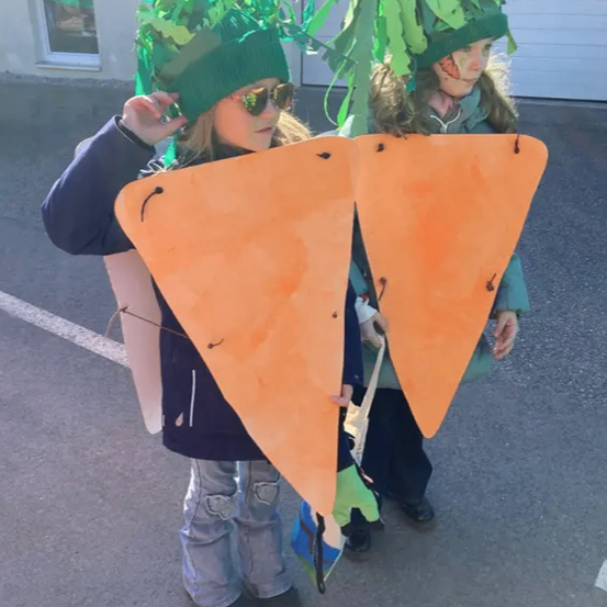 Zwei Kinder, verkleidet als Karotten, stehen draußen. Sie tragen grüne Mützen mit Blättern und halten orange Karottenausschnitte. Ein Gebäude mit einem Fenster ist hinter ihnen.