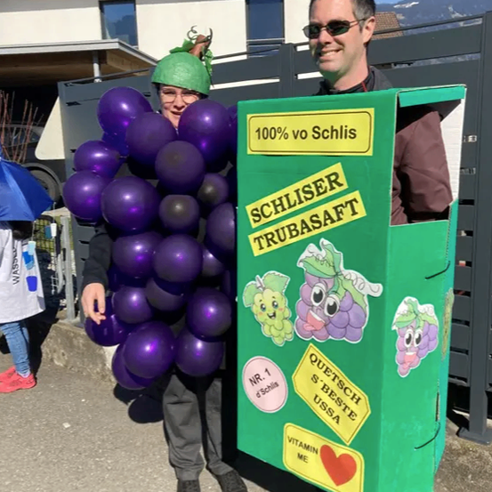 Zwei Personen in Kostümen stehen vor einem Gebäude mit Solarmodulen. Eine Person trägt ein Kostüm aus lila Luftballons und einer grünen Mütze. Die andere ist als Karton von Schliser Trubasaft, einem Traubensaft, verkleidet.