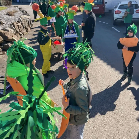 Eine Gruppe von Kindern in grünen Kostümen, einige mit karottenförmigen Masken, gehen auf der Straße. Im Hintergrund befinden sich Gebäude und geparkte Autos.