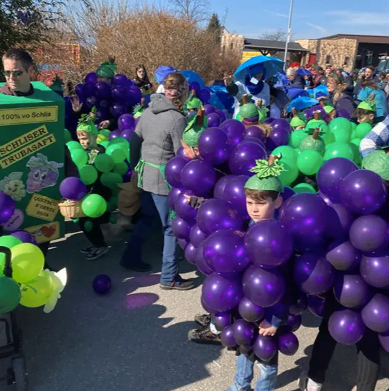 Eine Gruppe von Menschen in Kostümen, die Bündel von lila Luftballons halten, gehen während eines Umzugs auf einer Straße.