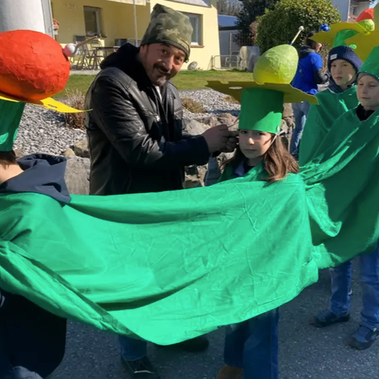 Ein Mann hilft einem Mädchen in einem grünen Kostüm, ein grünes Tuch zu halten. Sie sind draußen, mit Häusern und Menschen im Hintergrund.
