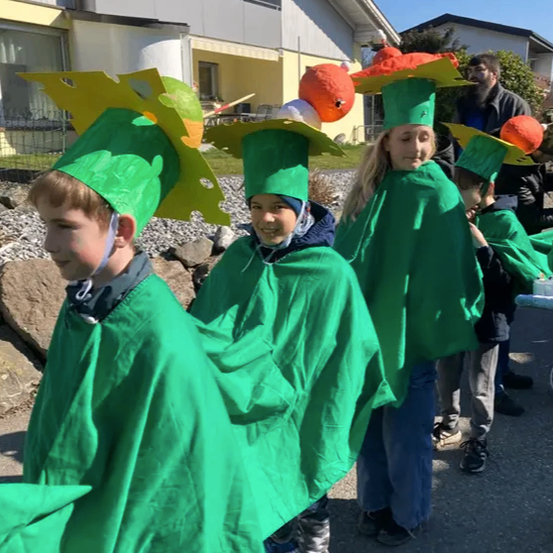 Kinder in grünen Kostümen und Papierhüten posieren vor einem Haus an einem sonnigen Tag. Dahinter beobachtet ein Mann, und eine große rote Papierblume ist sichtbar.