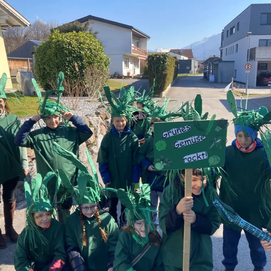 Eine Gruppe von Kindern in grünen Kostümen posiert für ein Foto auf der Straße, eines hält ein Schild mit der Aufschrift 'Grünes Gemüse rockt!'. Sie sind von Häusern und Grünpflanzen umgeben.