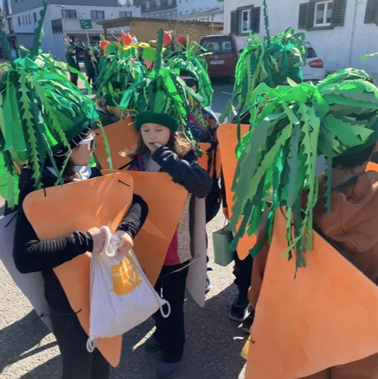 Eine Gruppe von Kindern, die als Karotten verkleidet sind, geht bei einer Parade. Sie tragen grüne Blattkronen und orangefarbene Karottenkostüme.