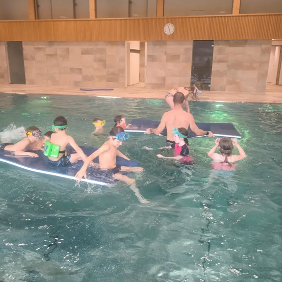 Kinder mit Schwimmbrillen und Schwimmhilfen treiben im Pool. Ein Mann hilft einem Mädchen im roten Badeanzug.