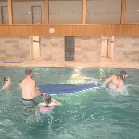 Eine Familie schwimmt in einem Pool mit einem blauen Surfbrett. Ein erwachsener Mann hält das Surfbrett, während Kinder darum herum spielen. Eine Uhr ist an der Wand über dem Pool angebracht.