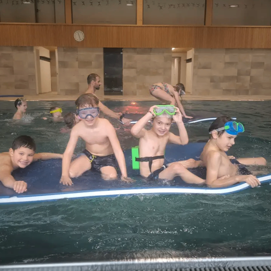 Mehrere Kinder mit Schwimmbrillen schwimmen auf einer aufblasbaren Matte im Pool. Eine Frau und ein Mann schwimmen im Hintergrund.