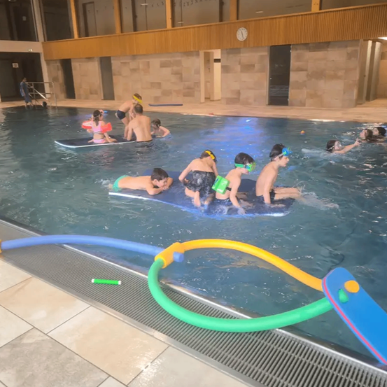 Eine Gruppe von Kindern in Schwimmbrillen und Schwimmhilfen schwimmt in einem Pool mit einer Person in der Nähe. Der Pool hat blaue und gelbe Wasserschläuche an der Seite.