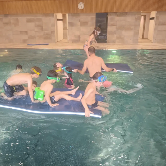 Mehrere Kinder schwimmen in einem Pool mit Schwimmbrillen. Ein Kind ist auf einem Schwimmbrett. Eine Frau geht in der Nähe des Pools.