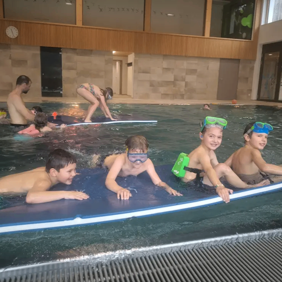 Kinder und Erwachsene schwimmen in einem Hallenbad, wobei eine Frau auf einem Surfbrett steht und andere darauf sitzen. Die Kinder tragen Schwimmbrillen.