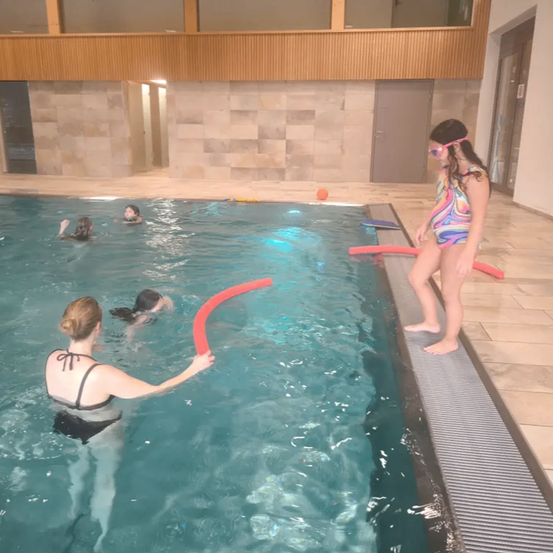 Eine Frau und mehrere Kinder schwimmen in einem Becken mit klarem blauem Wasser. Die Frau hält einen roten Schwimmreifen, während andere im Becken schwimmen. Ein Mädchen steht am Rand.