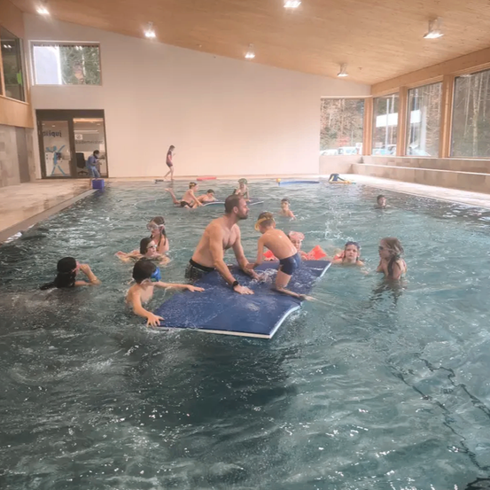 Ein Hallenbad mit mehreren schwimmenden Personen, darunter Kinder und Erwachsene, einige mit Schwimmbrillen. Ein Mann steht auf einem blauen aufblasbaren Schwimmbrett mit Kindern um ihn herum. Das Becken hat große Fenster und eine Holzdecke.