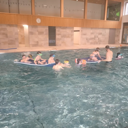 Eine Gruppe von Kindern in Schwimmbrillen und Badeanzügen schwimmt in einem Pool mit einem Trainer. Einige sind auf aufblasbaren Booten, andere schwimmen. Der Pool ist drinnen, mit Holzwänden und einer Uhr.