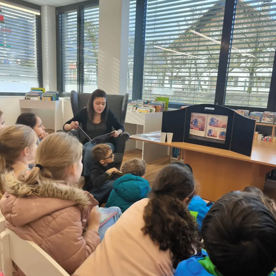 Eine Frau liest Kindern ein Buch vor, die in einem Klassenzimmer sitzen. Die Kinder sitzen aufmerksam vor ihr.