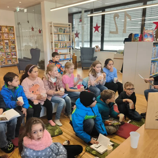 Eine Gruppe von Kindern sitzt in einem Kreis in einer Bibliothek, hält Bücher und Tassen. Der Raum hat Bücherregale, Stühle und Fenster mit Jalousien. Einige Kinder schauen zu einer Person, die in der Nähe der Bücherregale steht.