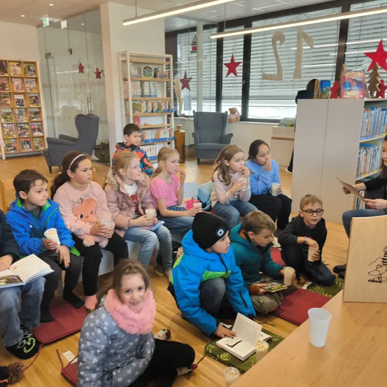 Eine Gruppe von Kindern sitzt auf dem Boden, einige mit Büchern, in einem Raum. Ein Kind liest ein Buch. Andere halten Tassen. Eine Frau steht und liest aus einem Buch.