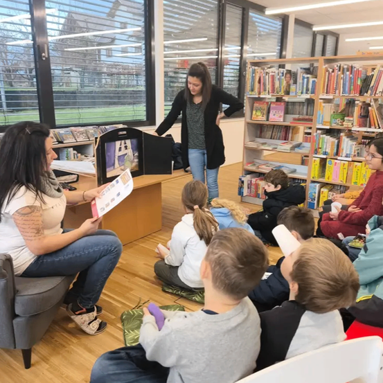 Eine Frau liest Kindern vor, die auf dem Boden in einer Bibliothek sitzen. Sie hält ein Buch, während eine andere Frau hinter ihr steht. Bücher stehen im Hintergrund in Regalen.