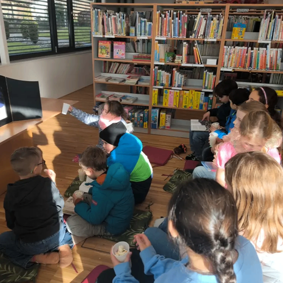 In einer Bibliothek sitzen Kinder auf dem Boden und lesen Bücher. Eine Person steht vor einem Regal voller Bücher. Der Raum hat einen Holzboden und Fenster.