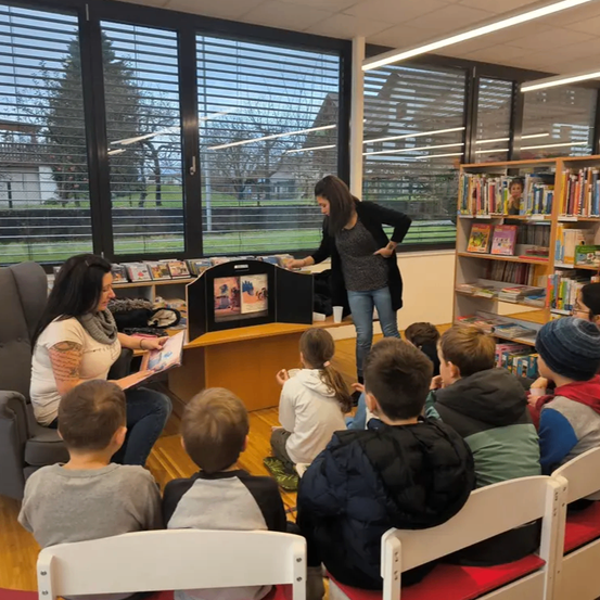 Zwei Frauen lesen Kindern in einer Bibliothek vor. Eine liest ein Buch, während eine andere hinter einem kleinen Bildschirm steht. Kinder sitzen auf Stühlen und hören aufmerksam zu.