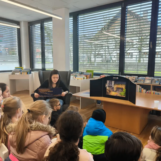 Eine Frau liest ein Buch vor einer Gruppe von Kindern, die vor ihr in einem Klassenzimmer sitzen. Das Klassenzimmer hat große Fenster mit Jalousien und einen Bücherregal mit Büchern.