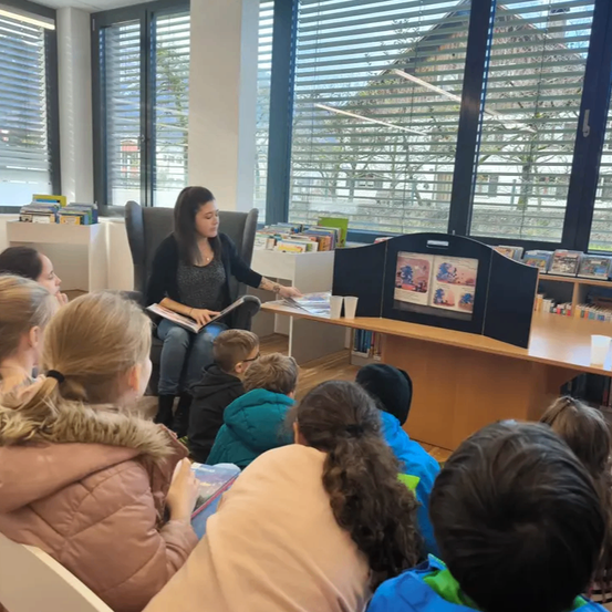 Eine Frau liest ein Buch für eine Gruppe von Kindern vor, die in Stühlen in einer Bibliothek sitzen. Der Raum hat große Fenster mit Jalousien, Bücherregale und einen Tisch mit einem Projektor.