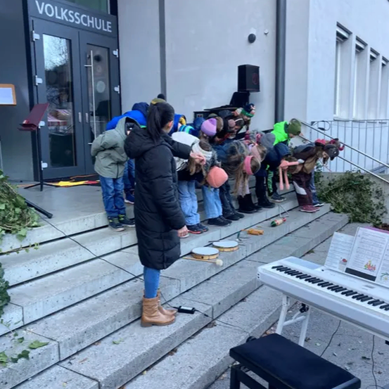 Eine Gruppe von Kindern in Winterkleidung spielt Musikinstrumente auf den Stufen vor einem Gebäude. Ein Klavier steht in der Nähe auf dem Boden.