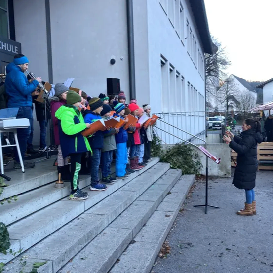 Eine Gruppe von Kindern mit Büchern singt auf den Stufen eines Gebäudes, begleitet von einem Mann, der Trompete spielt. Eine Frau in einem schwarzen Mantel steht neben einem Notenständer.