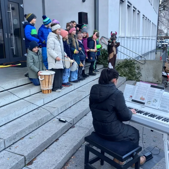 Eine Frau spielt Klavier auf einer Plattform, während mehrere Kinder und eine Frau in einem Kostüm zusehen. Ein Schlagzeuger steht in der Nähe.