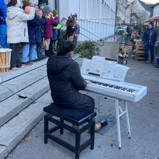 Eine Frau spielt Klavier auf der Straße vor einem Gebäude, während eine Menschenmenge zuschaut.