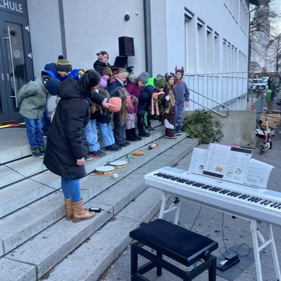 Eine Frau steht neben einem Klavier auf einer Treppe und unterrichtet eine Reihe von Kindern, die Trommeln halten. Die Kinder stehen vor einem Schulgebäude.