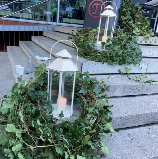 Zwei Laternen mit Kerzen und grünen Efeuranken auf Betonstufen, die zu einem Gebäude mit Glaswänden und einem Logo führen.