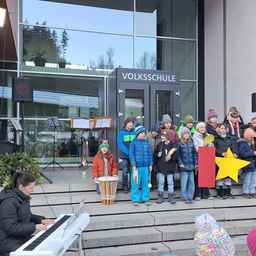 Eine Gruppe von Kindern, jedes mit Musikinstrumenten, steht auf Stufen vor einem Gebäude mit Glaswänden. Eine Frau spielt auf der linken Seite eine Tastatur. Das Gebäude ist mit 'Volksschule' beschriftet.