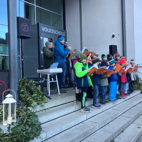 Ein Schulchor mit Erwachsenen und Kindern steht auf Stufen, hält Liederbücher und singt. Eine Person spielt Trompete. Das Volksschule-Schild ist über ihnen.