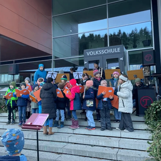 Ein Chor von Kindern in Winterkleidung steht auf Stufen vor einem Gebäude und hält orangefarbene Liederbücher. Das Gebäude hat eine Glaswand mit dem Schild 'Volksschule'.