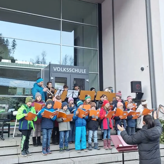 Ein Kinderchor singt vor einem Gebäude mit Glaswänden und einem Schild, das 'VOLKSSCHULE' besagt. Sie singen mit ihren Büchern und ein Dirigent leitet sie. Hinter dem Chor spielen Erwachsene Musikinstrumente. Die Szene ist draußen bei klarem Himmel.