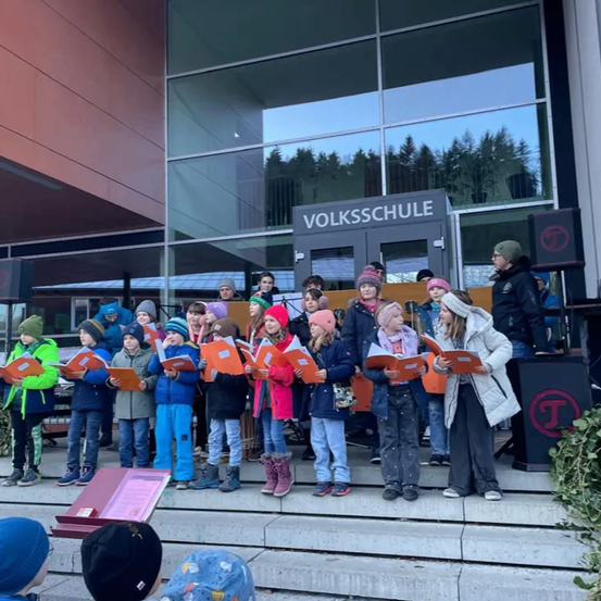 Eine Gruppe von Kindern, die orangefarbene Bücher halten, steht auf den Stufen vor dem Volksschule-Gebäude. Bäume sind durch die Glaswände sichtbar.