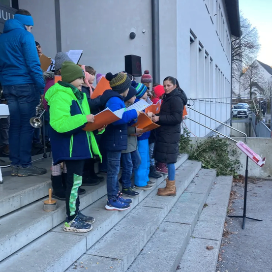 Eine Gruppe von Kindern steht auf Stufen und hält Musikbücher in der Hand, angeführt von einer Frau mit einem Mikrofon. Dahinter ist ein Gebäude, ein Baum und geparkte Autos zu sehen.