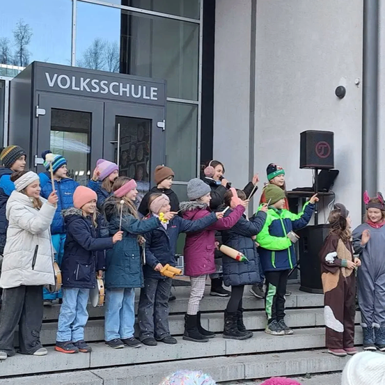 Kinder in Winterkleidung stehen auf den Stufen einer Schule und halten Musikinstrumente in der Hand. Über der Tür steht der Name der Schule, 'VOLKSSCHULE'.