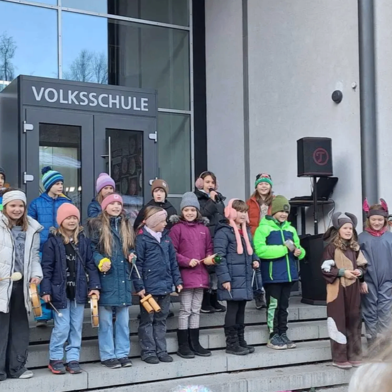 Eine Gruppe von Kindern in Winterkleidung posiert auf den Stufen vor einem Schulgebäude. Einige halten Musikinstrumente und eine Person steht hinter einem Lautsprecher.