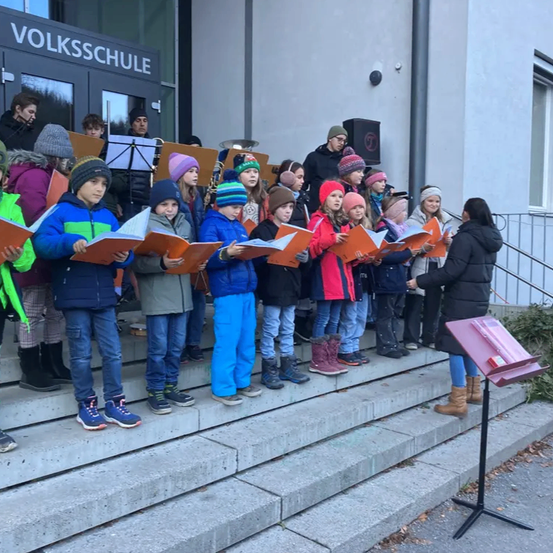 Ein Kinderchor in Winterkleidung steht auf Stufen vor einem Gebäude, hält Notenbücher und singt.
